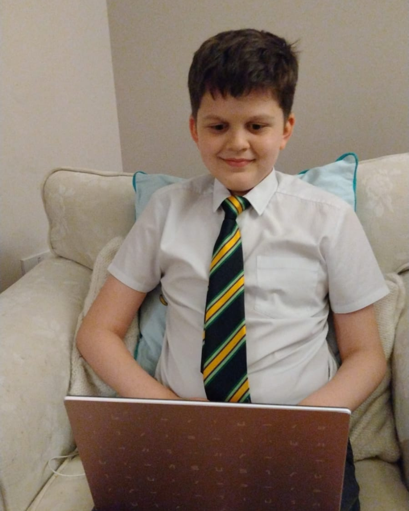 Joshua is sat on an armchair with his laptop on his knee. He has short dark hair, is wearing his school shirt and tie, and is smiling.