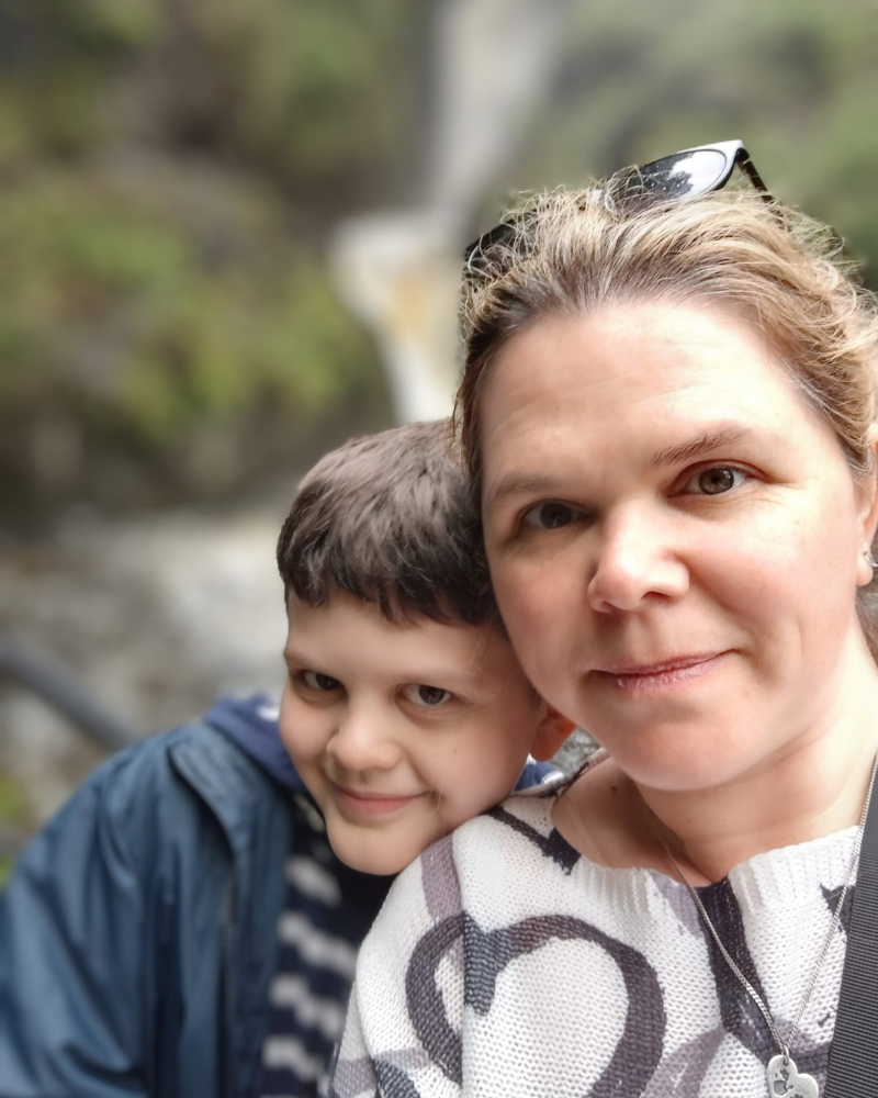 Joshua and his mum are pictured with a blurred waterfall behind them. As mum takes the selfie. Joshua rests his head on her shoulder. They are both smiling.