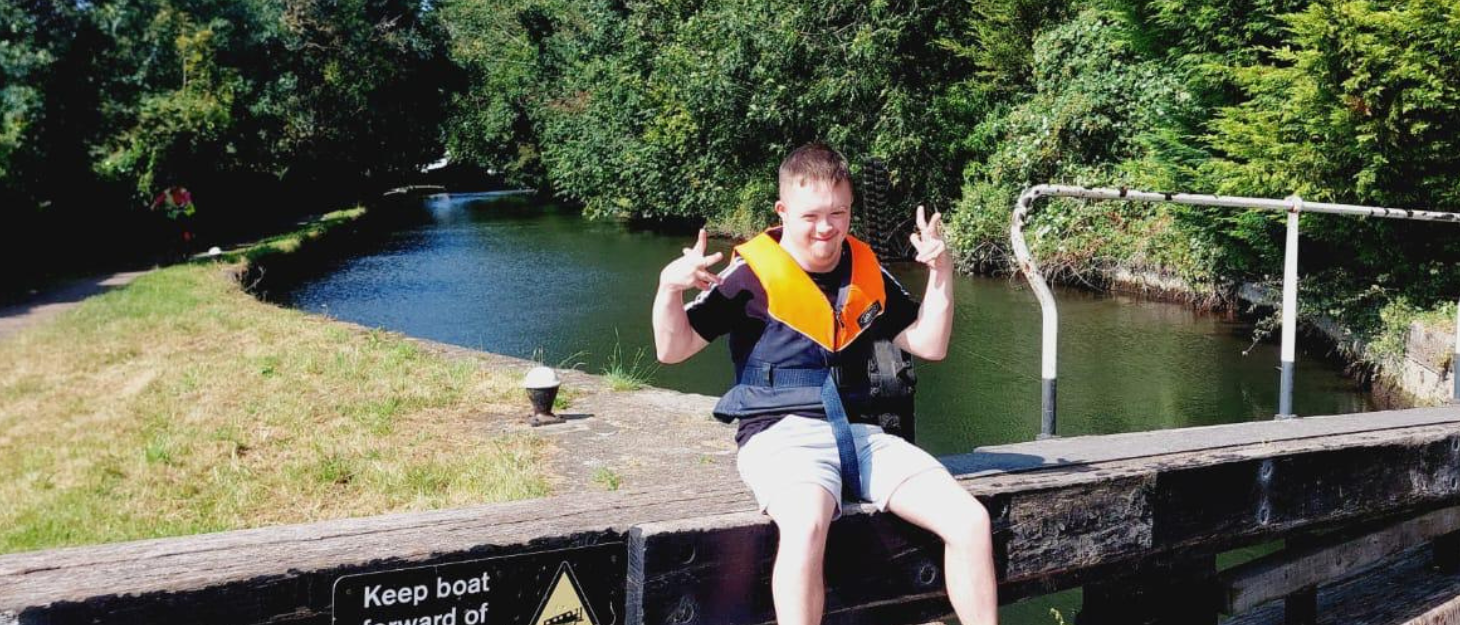 Josh is sat by the canal. He wears a life vest and he is posing using his hands for the camera. It is a sunny day so he wears shorts and a t-shirt.
