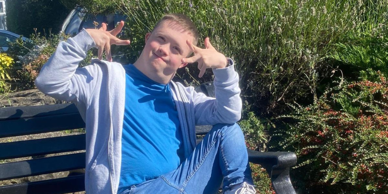 Josh is sat on a park bench and he is grinning, holding both hands in almost 'peace' like signs. He is wearing a blue t-shirt and a grey zip hoodie.