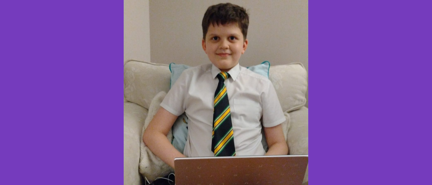 Joshua is wearing his school shirt and tie and is smiling. He is sat on an armchair with his laptop on his knee, likely creating away on Canva. He has short dark hair.
