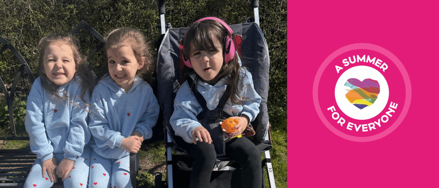 Young girls triplets sat in the sun together, one child is in a pushchair and wearing pink ear defenders