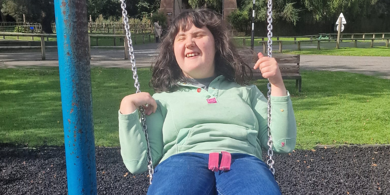 Kirsten is smiling whilst sat on a swing in a park. She is holding onto the chains and leaning back.