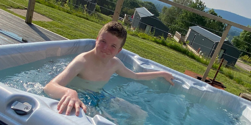 Lennon is sat in a hot tub on a sunny day with greenery behind him. He is holding onto the sides of the hot tub.