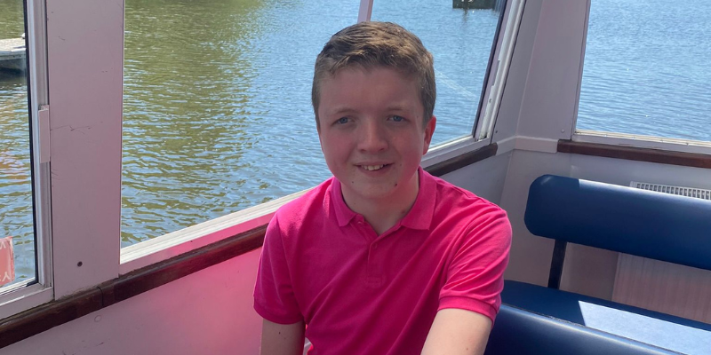 Lennon is wearing a bright pink shirt and is sat on a boat. He is smiling.