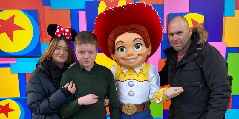 Lennon and his parents are stood with Jessie from Toy Story. Lennon's mum wears Minnie Mouse ears, and they are all in front of a bright background for taking photos.