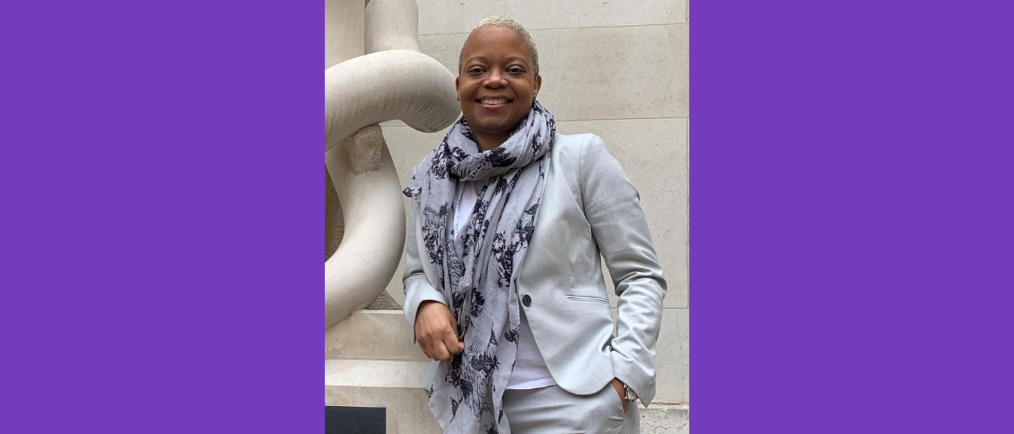 A lady wearing a light grey suit with a matching scarf leaning against a sculpture.
