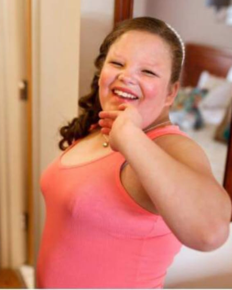 Melissa is wearing her brown hair curled to her right side, and a pink vest. She has her left hand up to her chin and is smiling, with rosey cheeks.