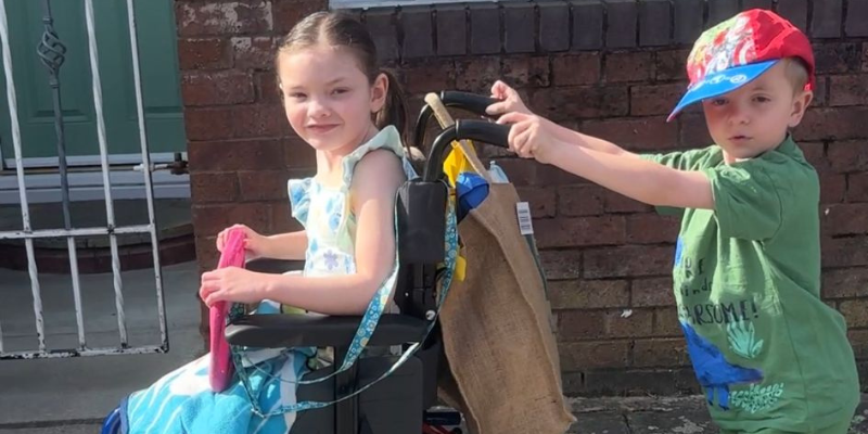 Matthew is pushing his sister Olivia in her wheelchair on a sunny day. Olivia is wearing a dress and Matthew a sun cap. Olivia is smiling.