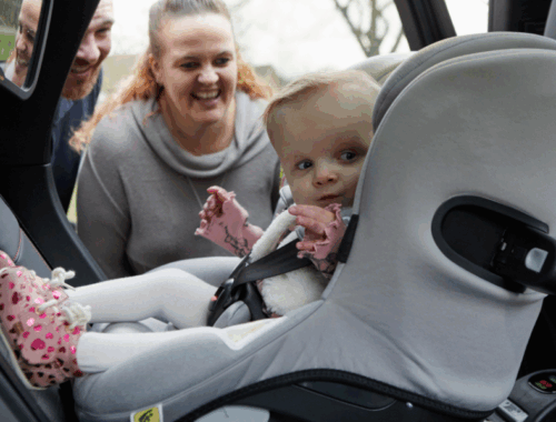 Olivia sits in her car seat in the back of their Family Fund Motability Programme lease car and her mum and dad smile at her through the open car door.