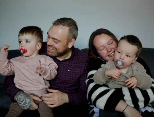 A family sitting on the sofa cuddling together. Dad holding the youngest son and mum holding the two year old son