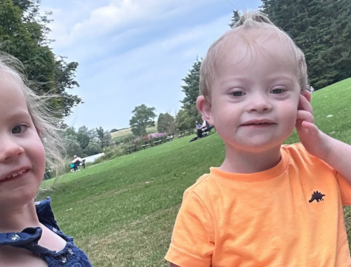 Parker, a young boy with Down Syndrome, sits in a park with his older sister. They are smiling at the camera.