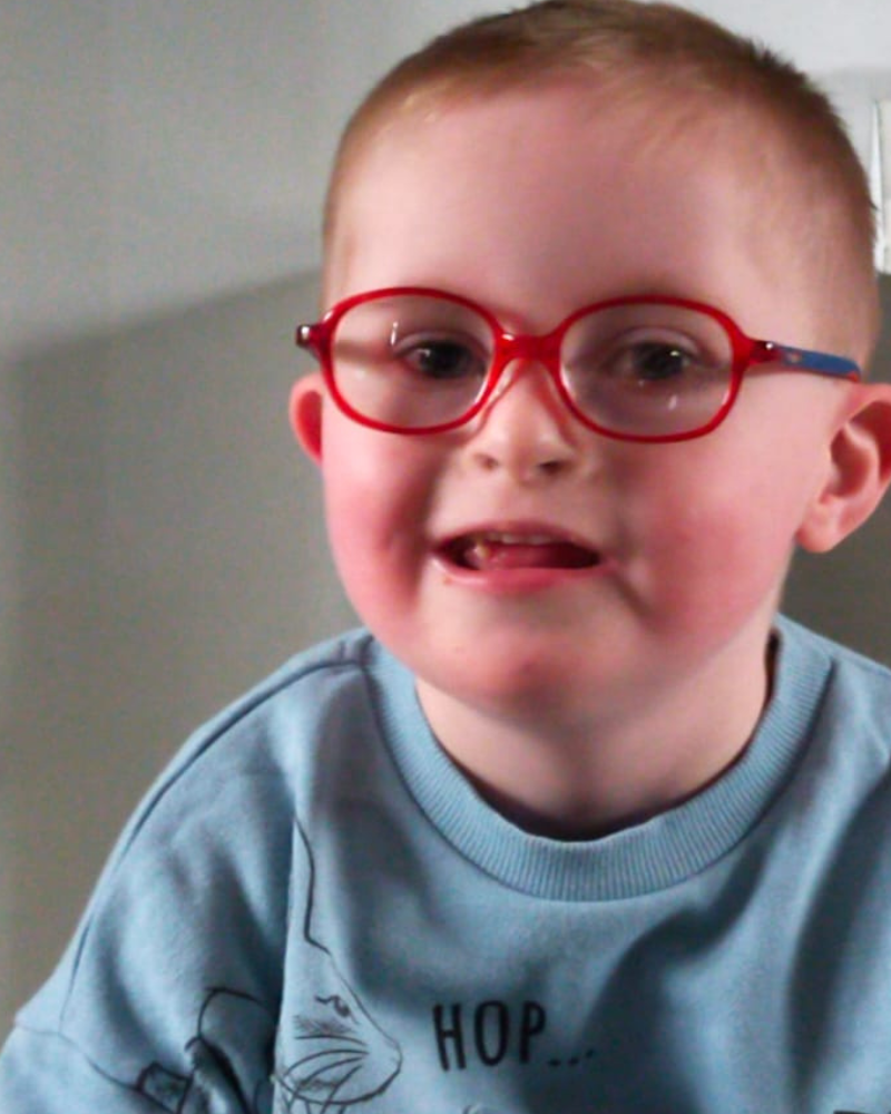 Lewis is smiling, wearing red glasses with rosey cheeks. He has short hair and wears a blue jumper.