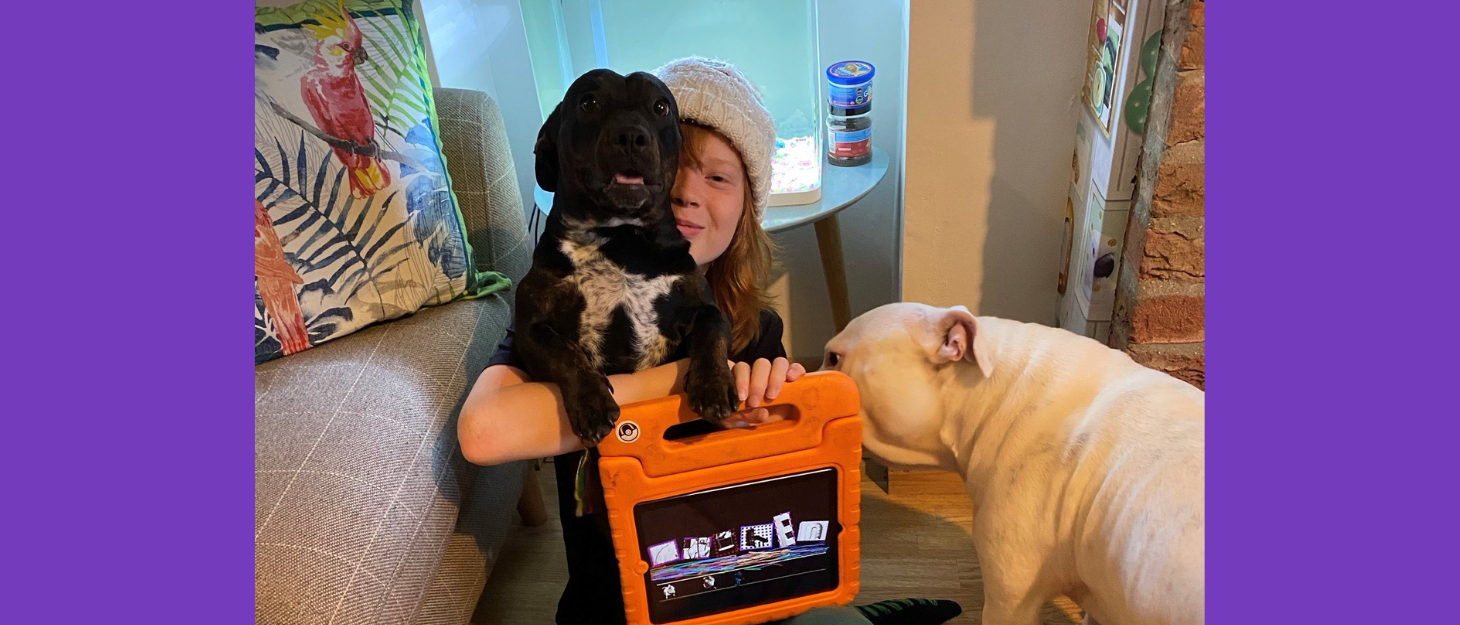 Boy wearing hat cuddles a dog in one hand and holds up his orange iPad in the other