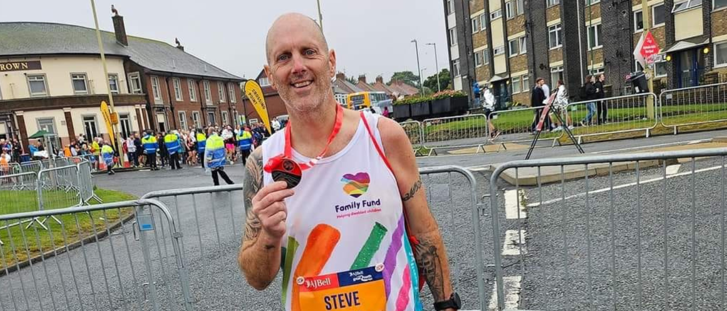 Steve is stood at The Great North Run holding his medal in one hand proudly. He is wearing his Family Fund running vest and a running number, and is smiling.