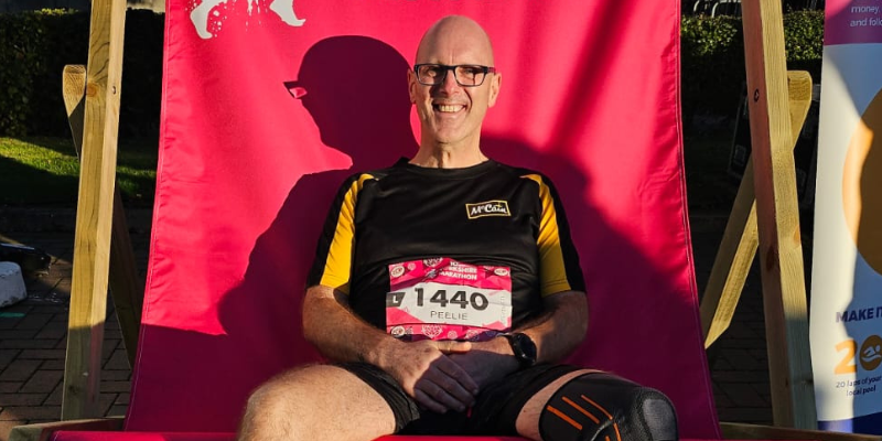 Steve is sat in the last of the day's sunshine on a very large sunlounger, likely at an event. He is wearing running gear and a number, and he has just taken part in a running event. He is wearing glasses and smiling.