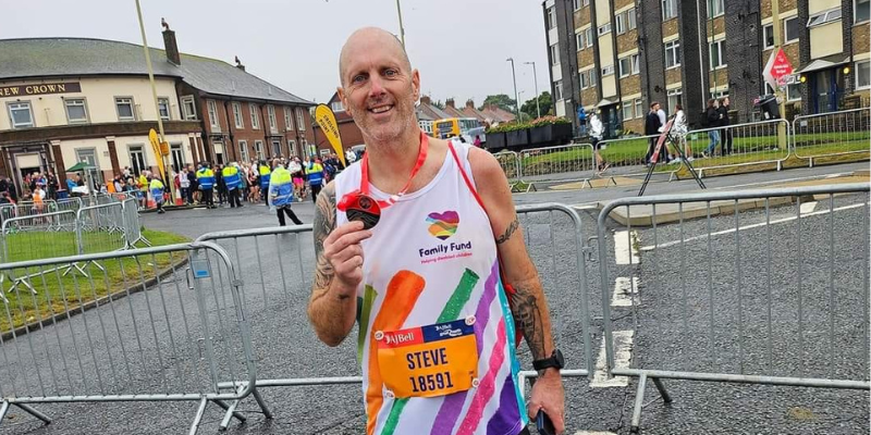 Steve is stood at The Great North Run holding his medal in one hand proudly. He is wearing his Family Fund running vest and a running number, and is smiling.