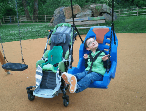 A young boy in a specialised swing next to a buggy in a park.