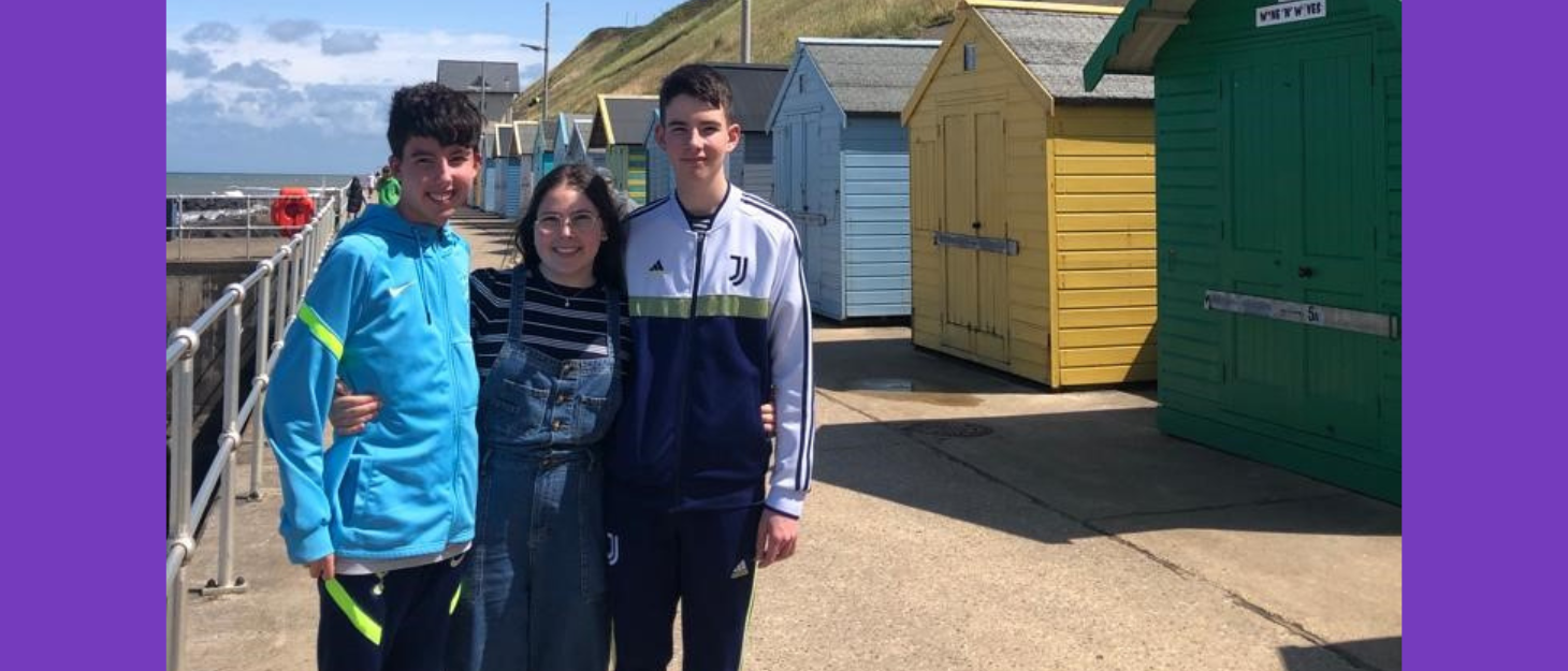Three family memebers stood at the seaside by some colourful beach huts