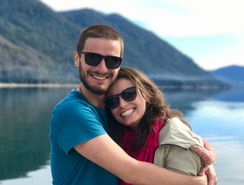 A couple wearing sunglasses, standing by a loch smile into the camera. Take a Break Scotland is open now.