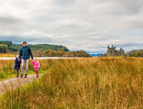 A dad walking with two small children across a field in Scotland with a castle in the background