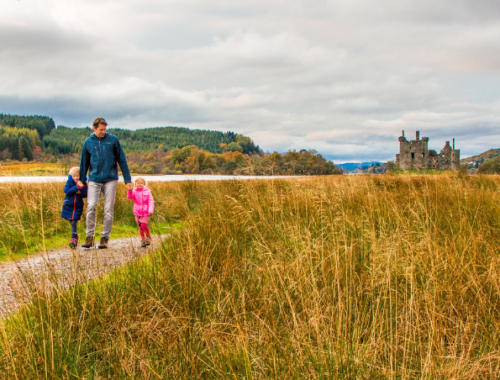 A dad walking with two small children across a field in Scotland with a castle in the background