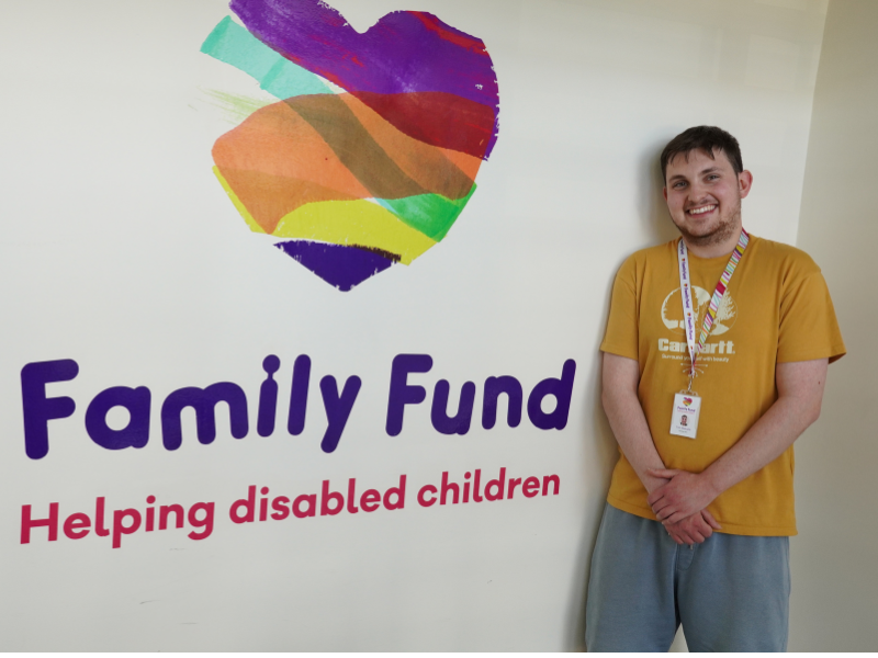 A young male intern wearing a yellow t-shirt is smiling and standing next to the Family Fund logo on a wall in an office