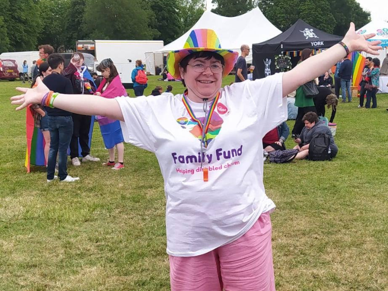 Tracey stands at York Pride wearing a rainbow hat and a Family Fund t-shirt. She's smiling and raising her hands in the air.