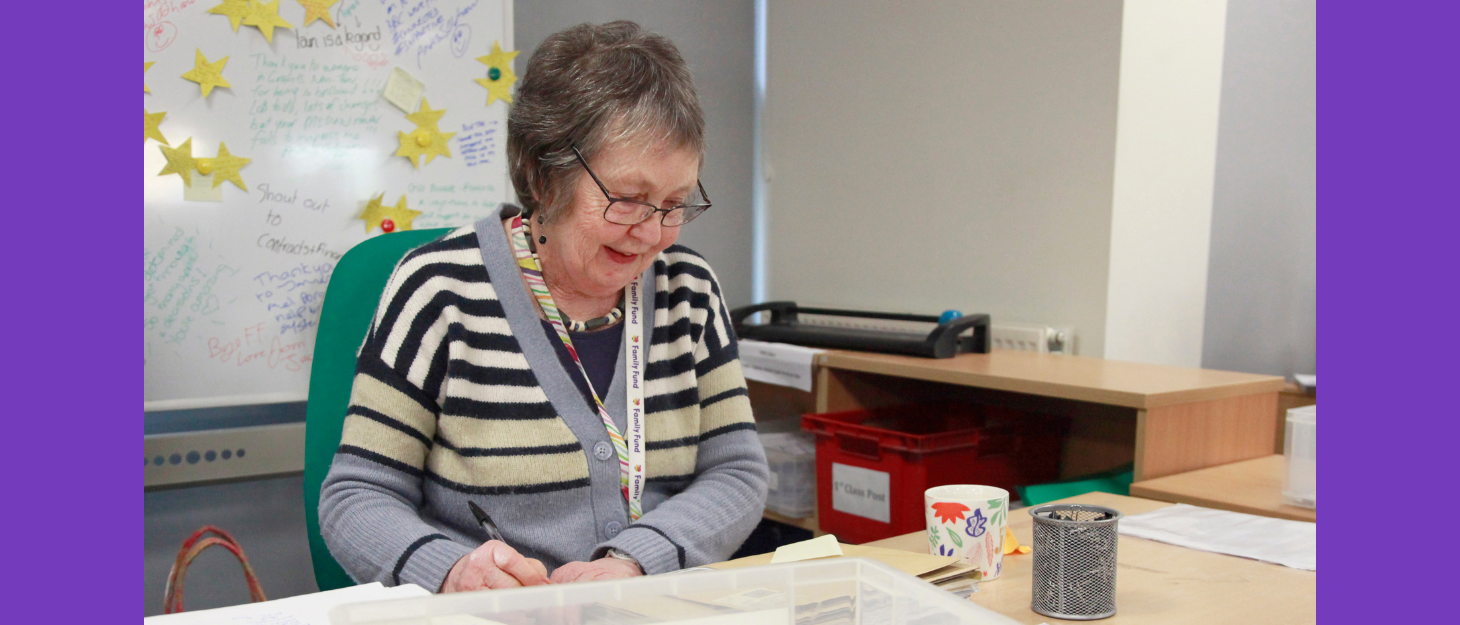 A woman wearing a family fund lanyard and a striped shirt is sitting at a desk, sorting mail and smiling